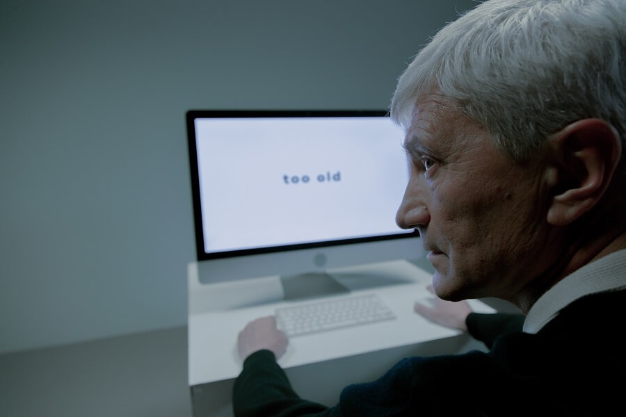 An Elderly Man Sitting Beside the Computer with a sign that says he is too old for the job
